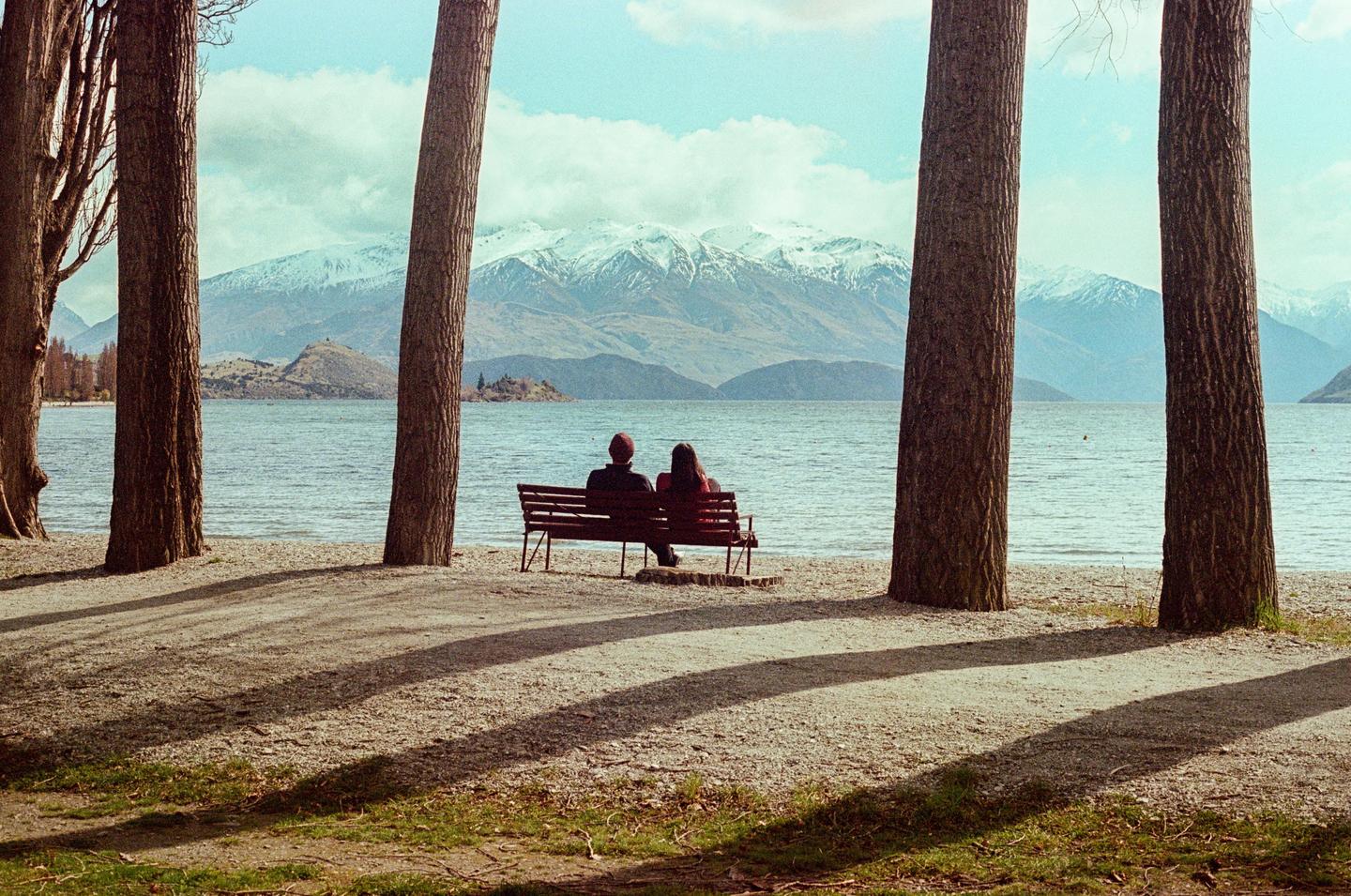A couple sits on a bench by a lake with snow-capped mountains in the background, framed by tree trunks.