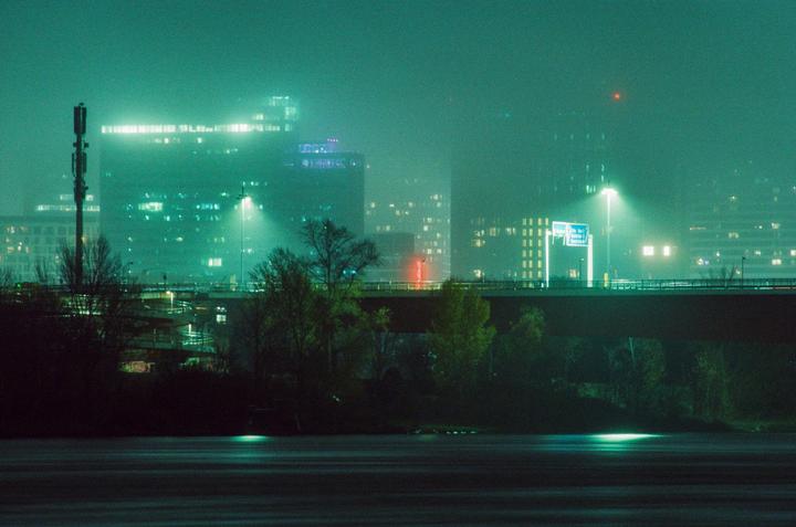 A city skyline with illuminated buildings and a bridge is visible through a thick fog at night.
