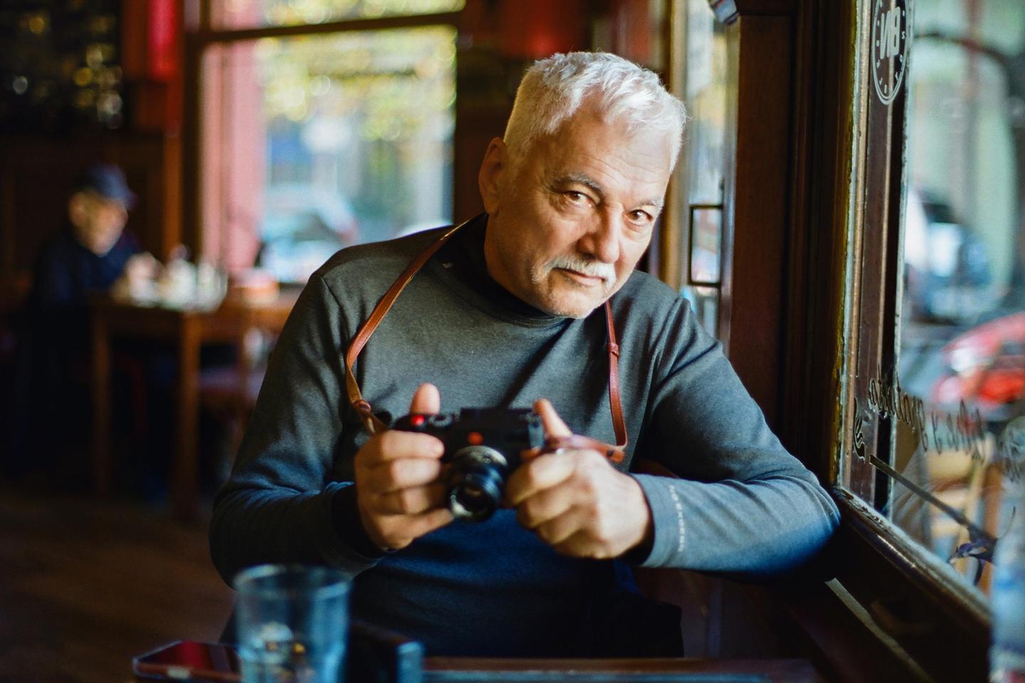A man with a mustache and white hair holds a camera while sitting by a window in a cafe.