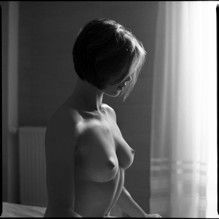 A woman with short hair stands topless in front of a window with a curtain.