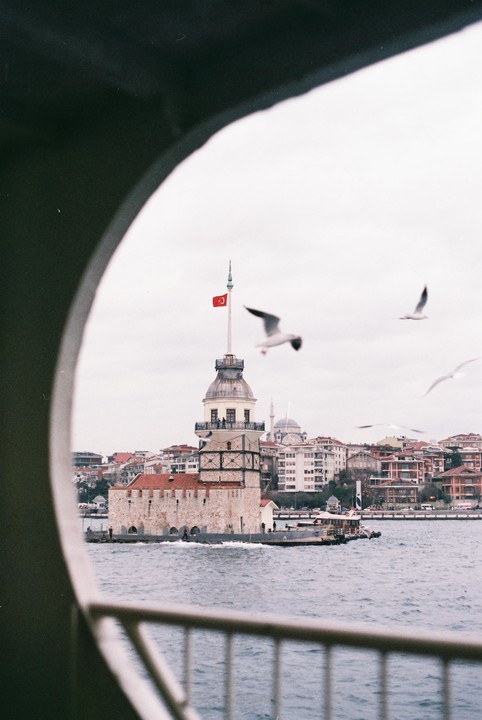 The Maiden's Tower with a Turkish flag is visible from a boat, with seagulls flying around it and a city in the background.