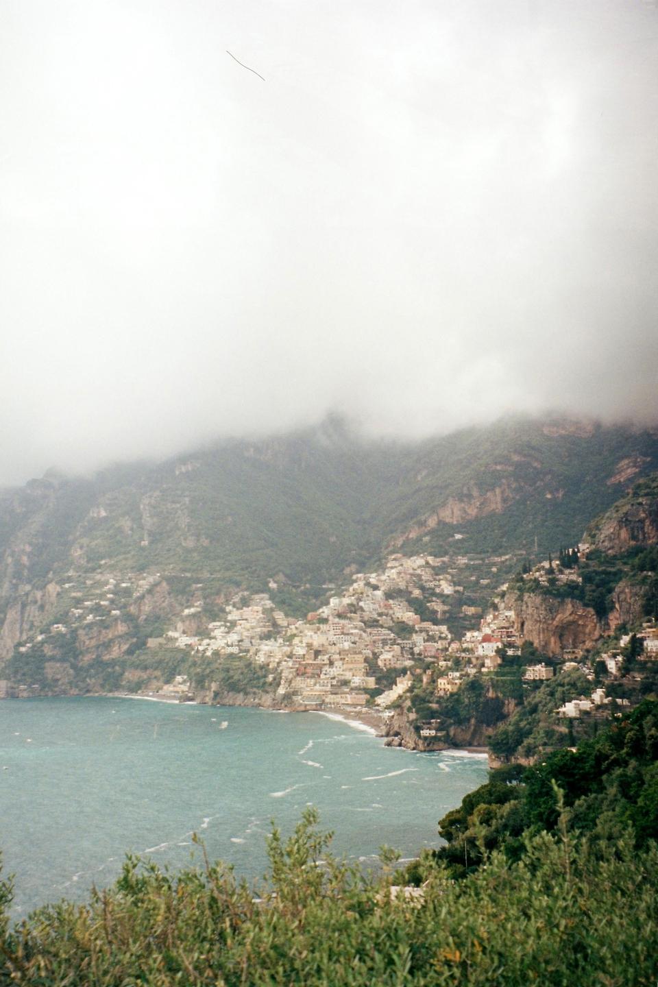 A coastal town with many buildings is nestled into a steep, green mountainside under a cloudy sky.
