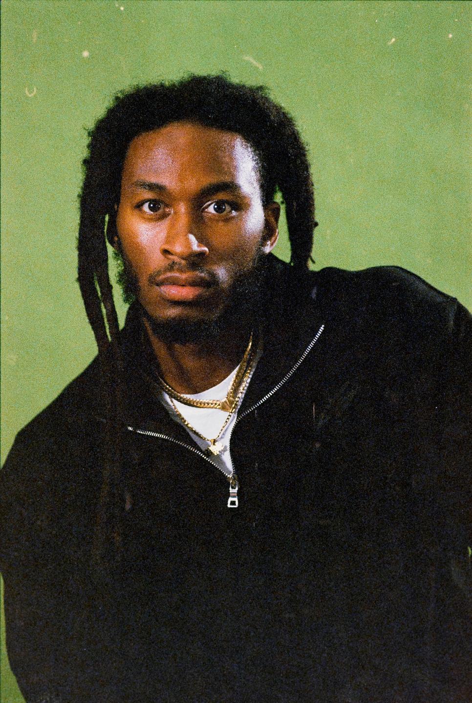 A man with dreadlocks and a beard wears a black jacket and gold chains against a green background.
