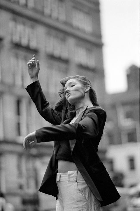A woman with her eyes closed and arms raised dances in front of a building.