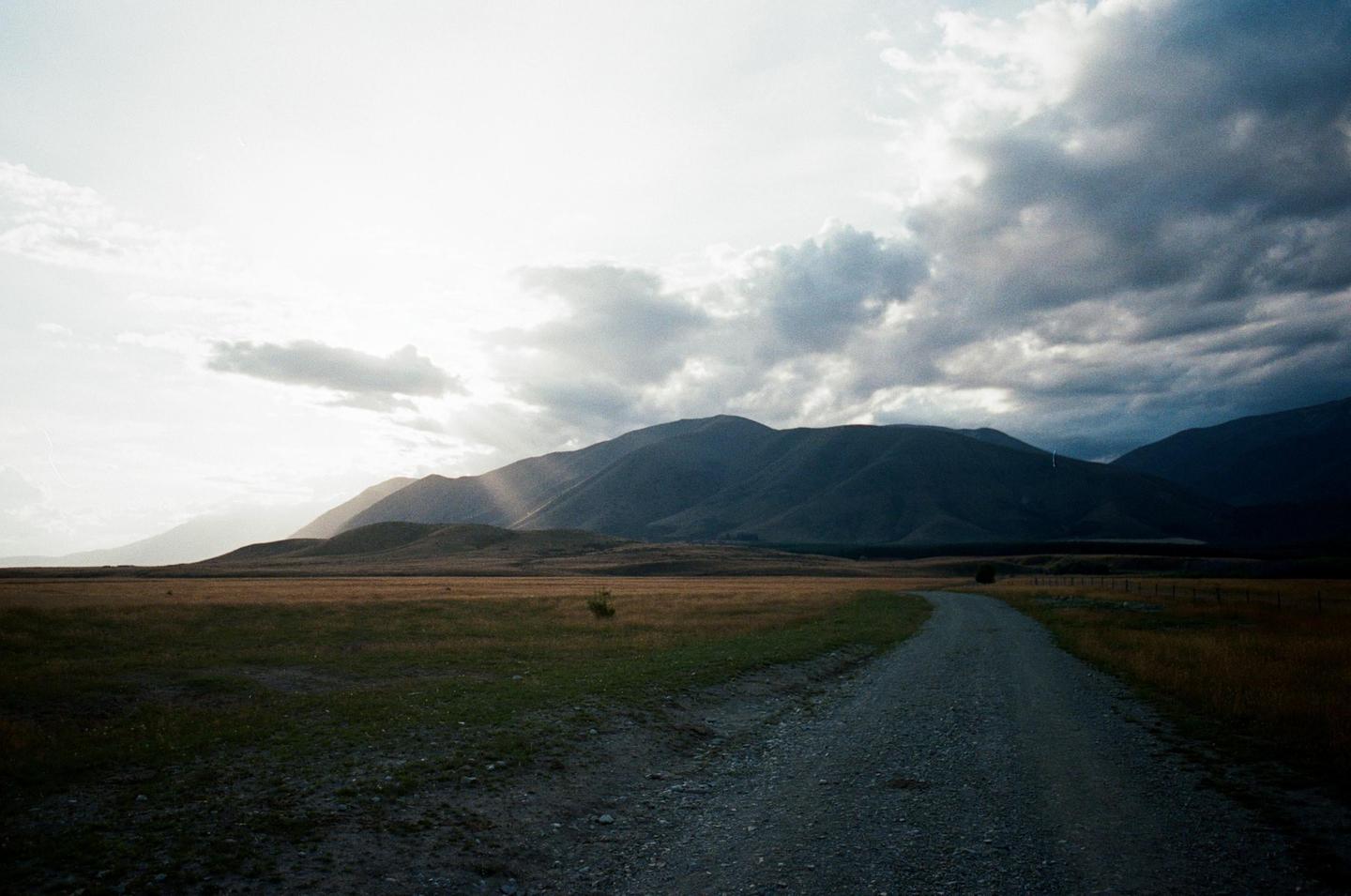 A gravel road leads through a field towards mountains under a cloudy sky with sun rays breaking through.