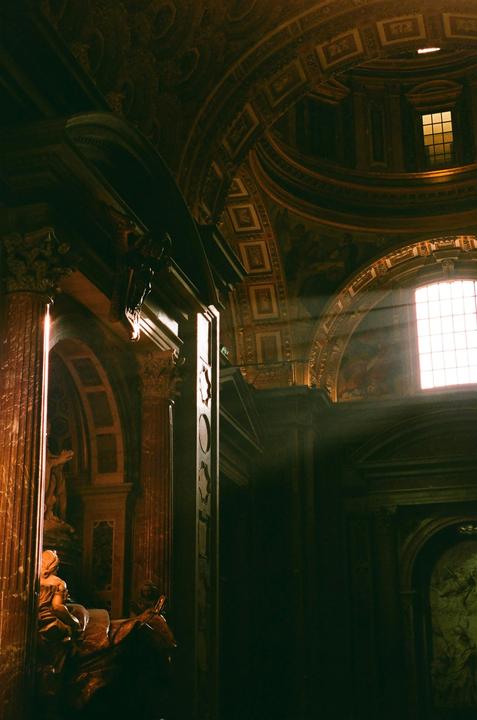 Sunlight streams through a large window into a richly decorated, dark interior with a vaulted ceiling and ornate columns.