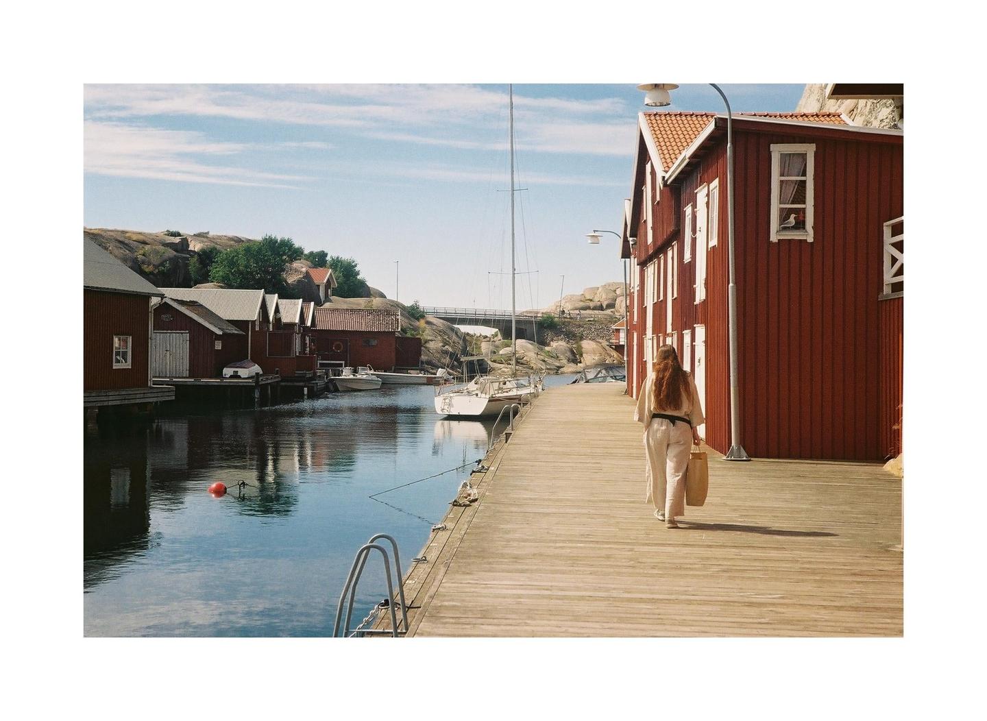 A woman walks along a wooden pier next to a canal with red buildings and boats.