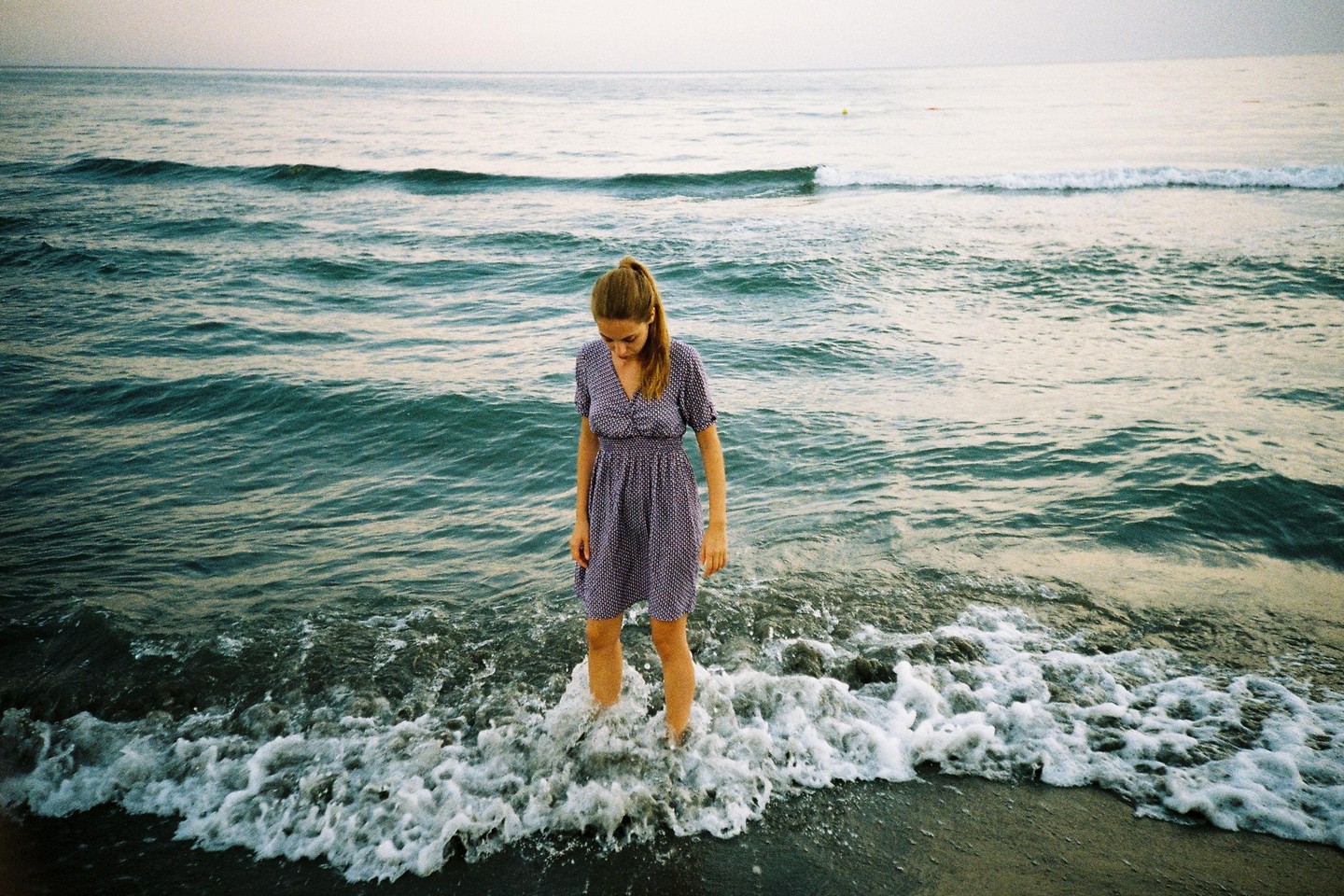 A woman stands in the shallow water at the edge of the ocean, looking down at the waves.