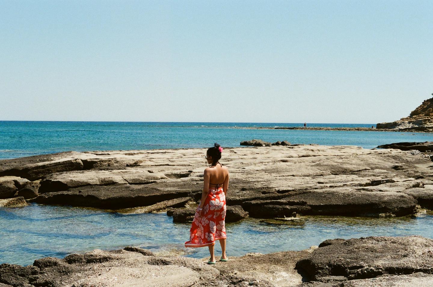 A woman stands on a rocky shore looking out at the ocean under a clear sky.