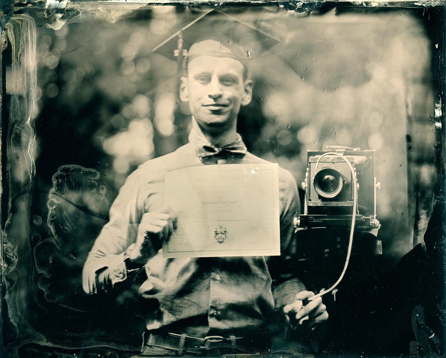 A man in a graduation cap and bow tie holds a diploma and a large format camera.
