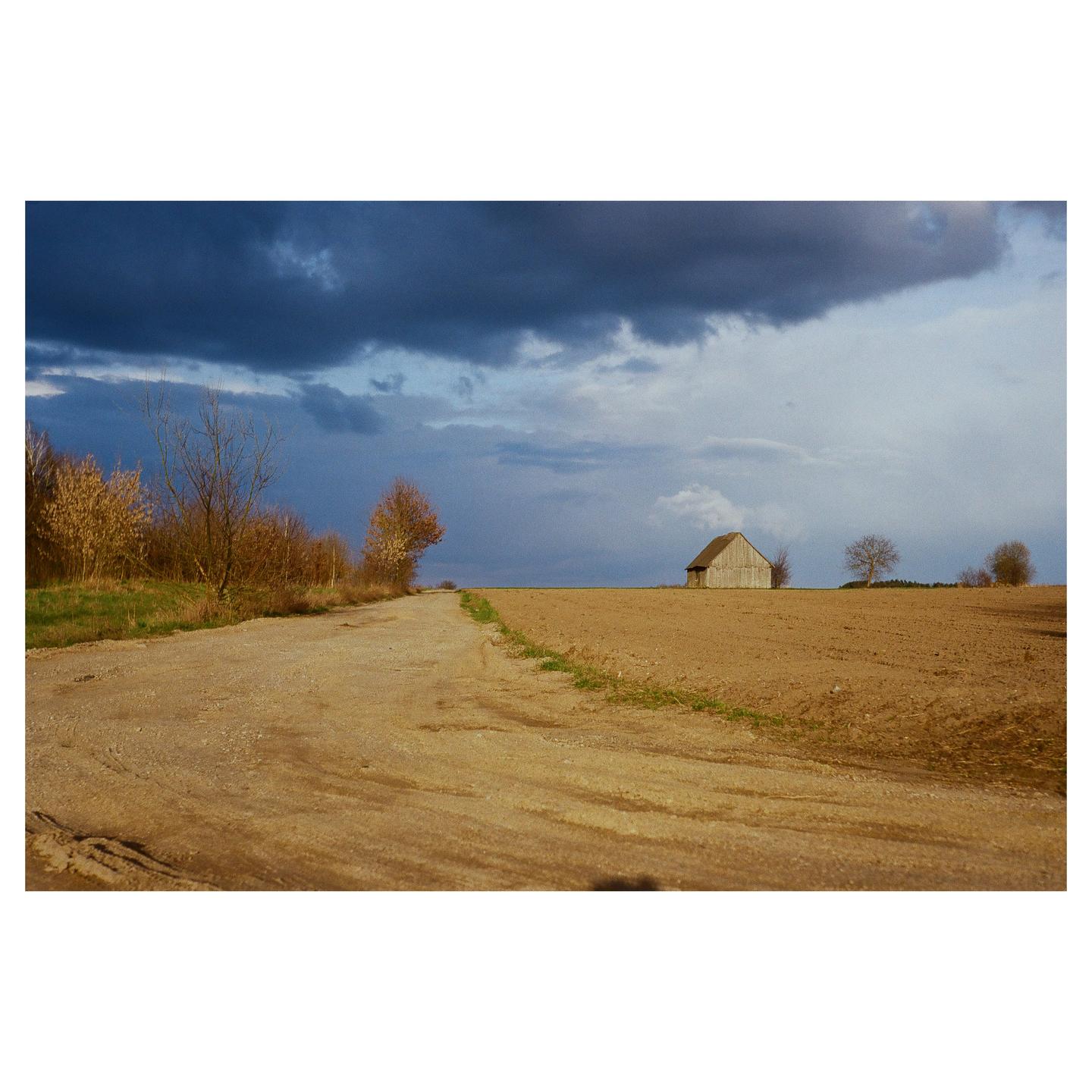 A dirt road leads through a field towards a small wooden barn under a dramatic sky.
