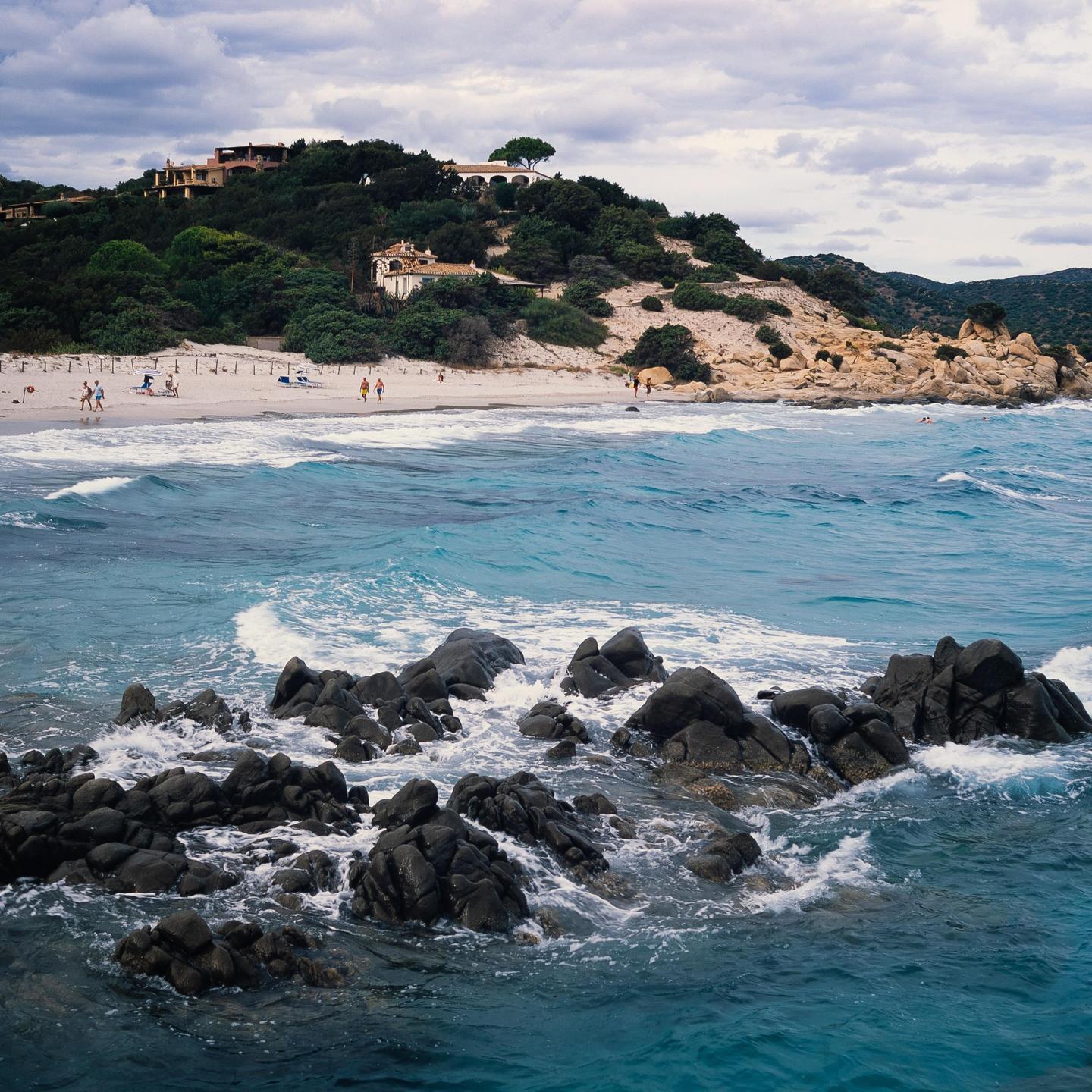 A rocky coastline with turquoise water and a sandy beach is visible under a cloudy sky.