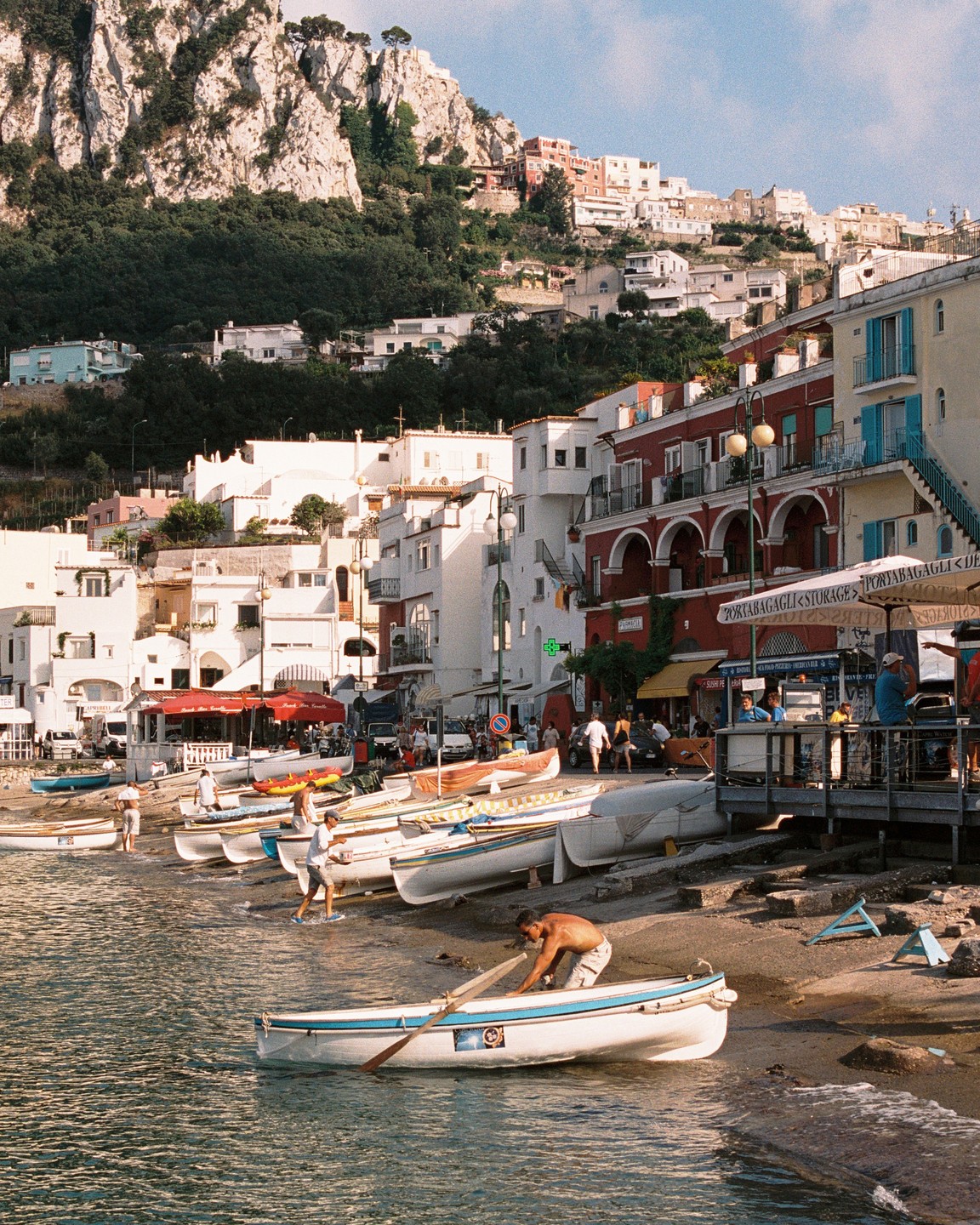 A man pushes a rowboat into the water on a beach lined with boats, with a town built into a hillside in the background.