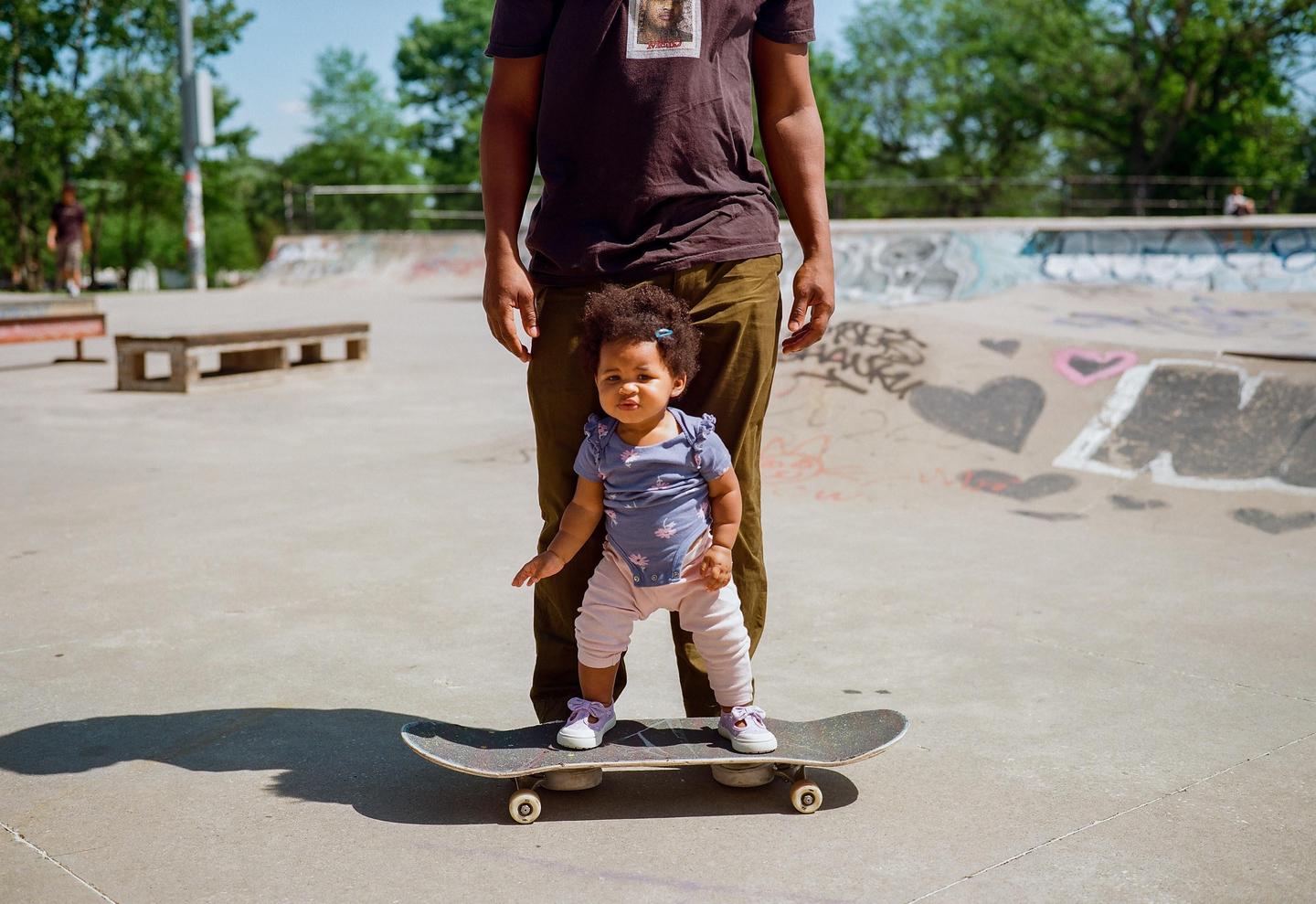 A baby stands on a skateboard in a skatepark with an adult standing behind them.