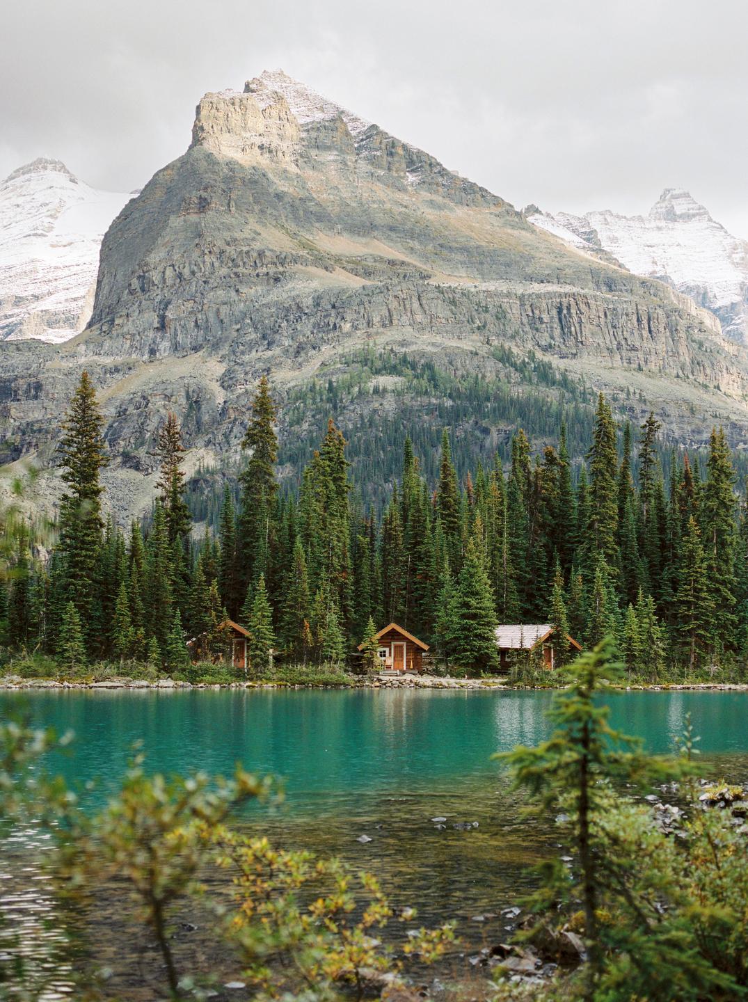 Two small cabins sit at the edge of a turquoise lake, backed by a dense forest and a large mountain.