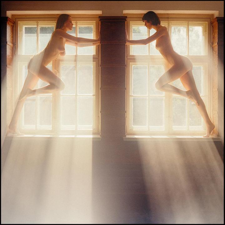 Two women pose nude on window sills, illuminated by rays of sunlight streaming through the windows.