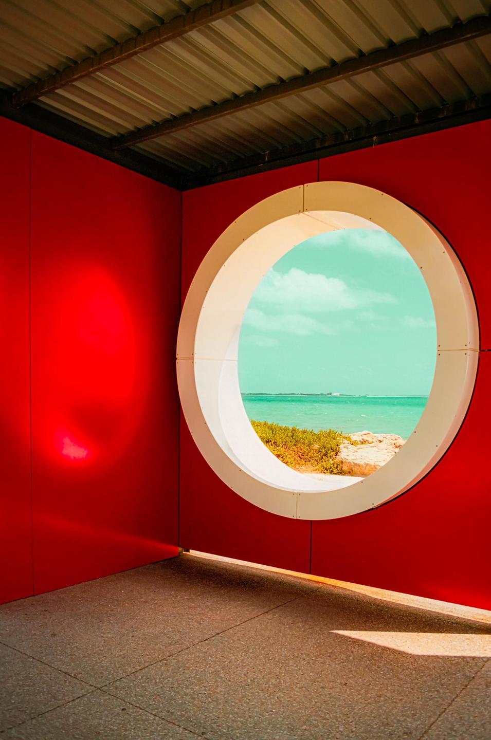 A circular window in a red wall looks out onto a bright blue ocean and sky.
