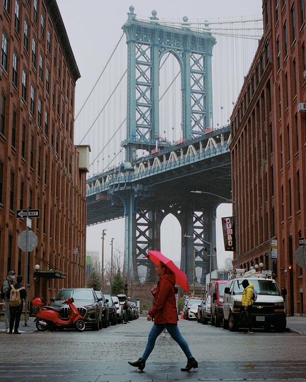 A woman with a red umbrella crosses a street with the Manhattan Bridge in the background.