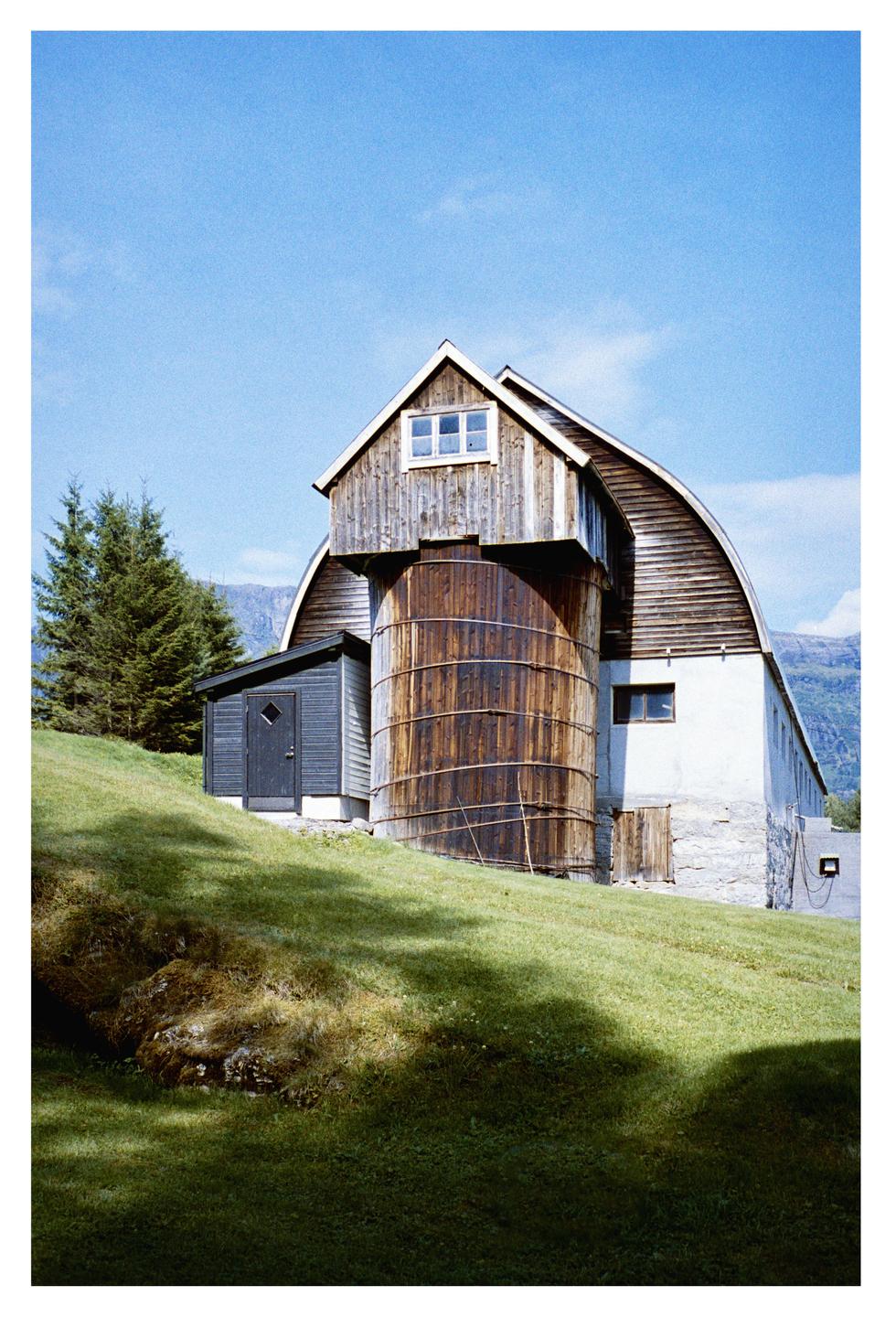 A barn with a large wooden silo attached to its side stands on a grassy hill under a clear blue sky.
