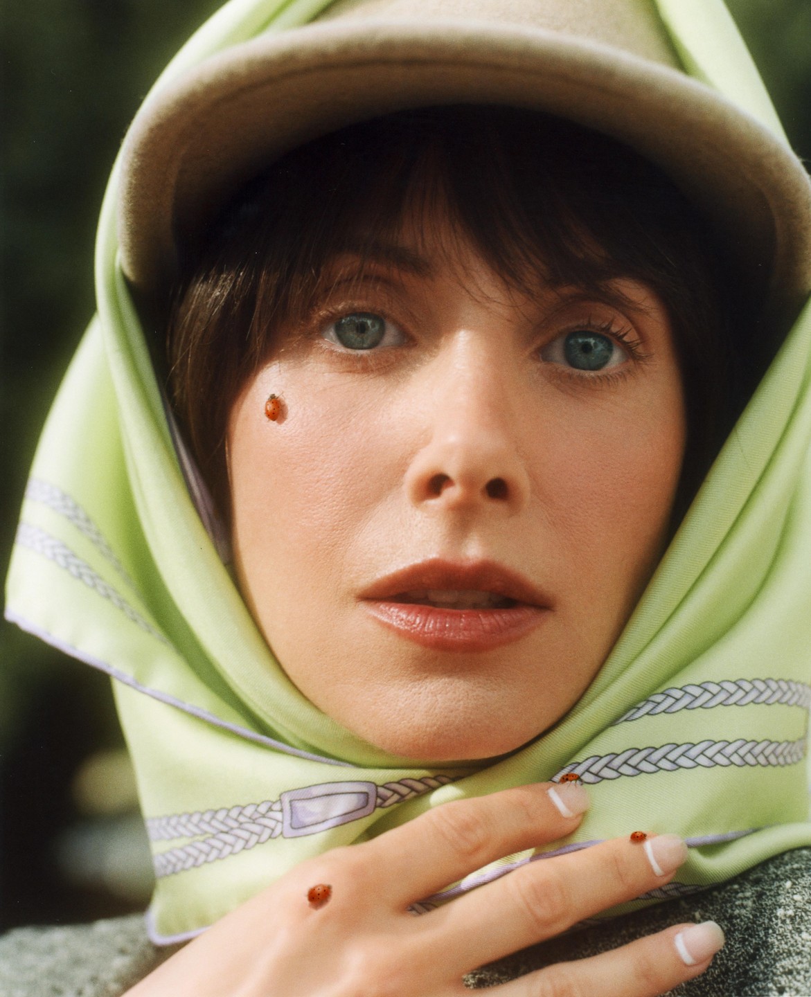 A woman with a green headscarf and a hat looks forward with ladybugs on her face and hand.