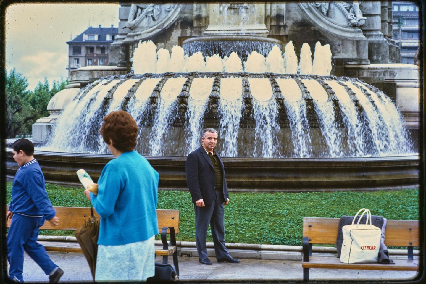 A man stands in front of a large fountain while a woman and boy walk past in the foreground.