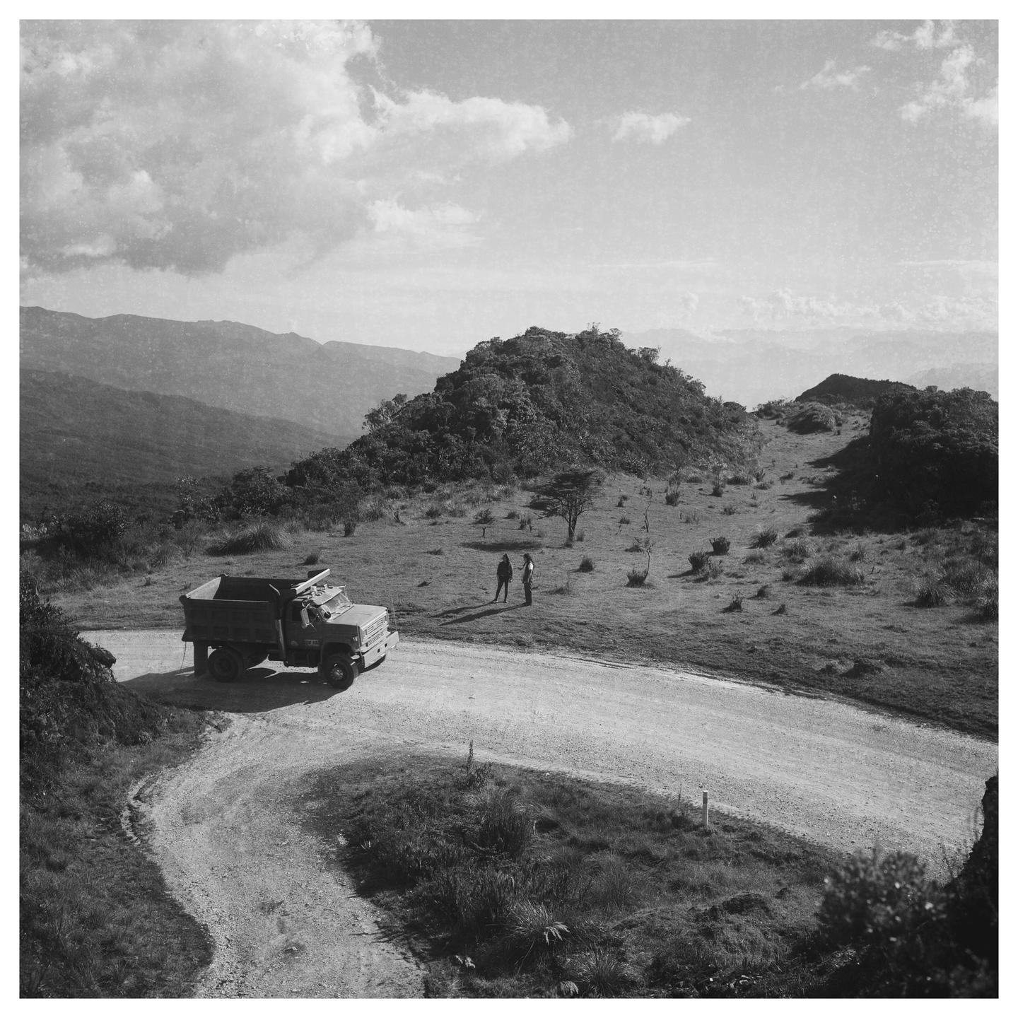 A dump truck is parked on a winding dirt road with two people standing nearby, surrounded by hills and mountains under a cloudy sky.
