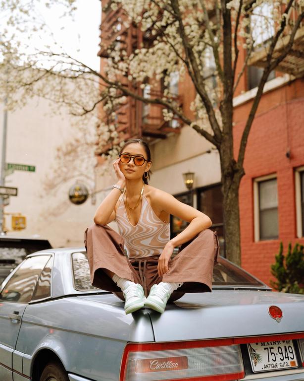 A woman wearing sunglasses and a halter top sits on the trunk of a car on a city street.