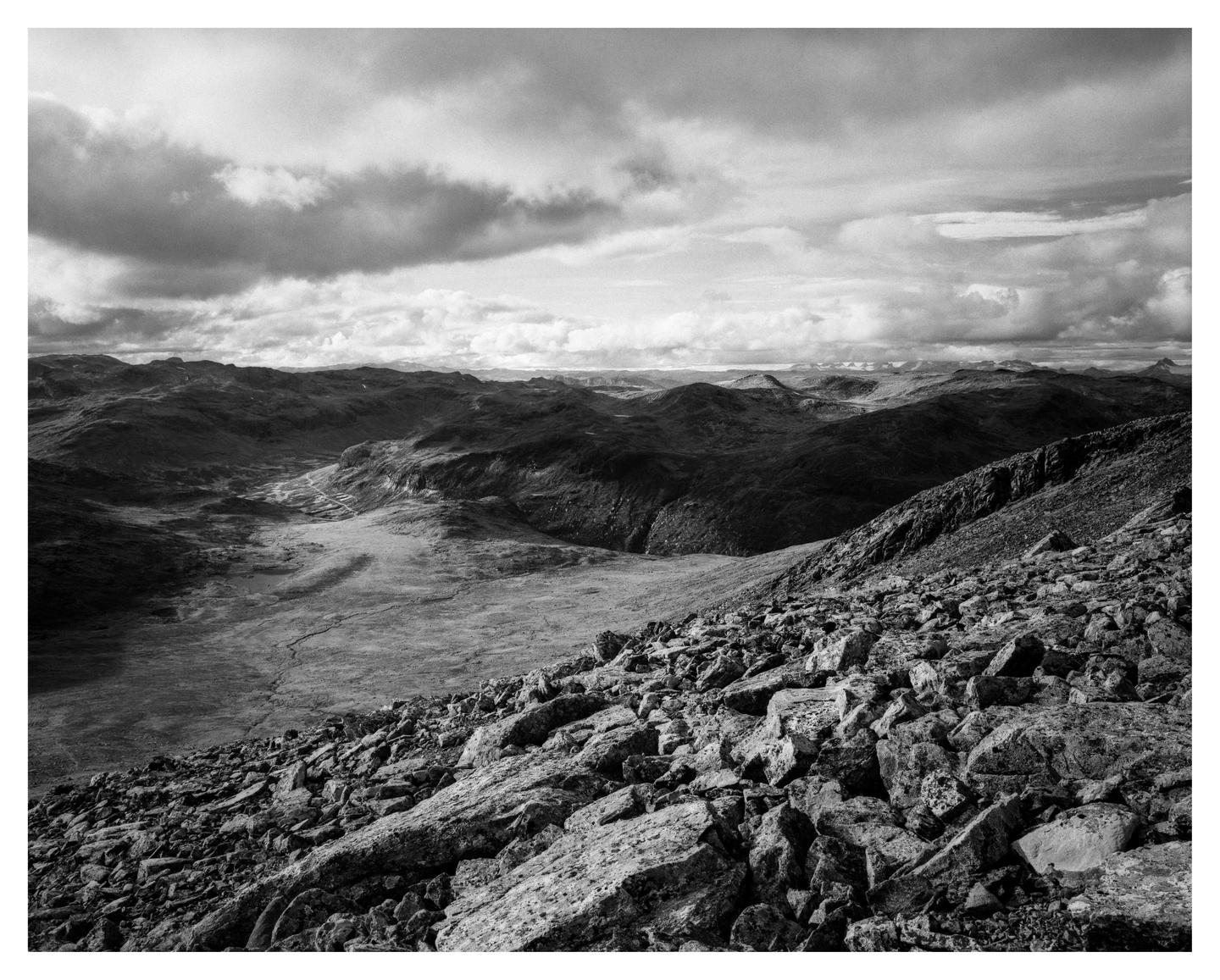A black and white landscape shows a rocky slope in the foreground leading to a valley and distant mountains under a cloudy sky.