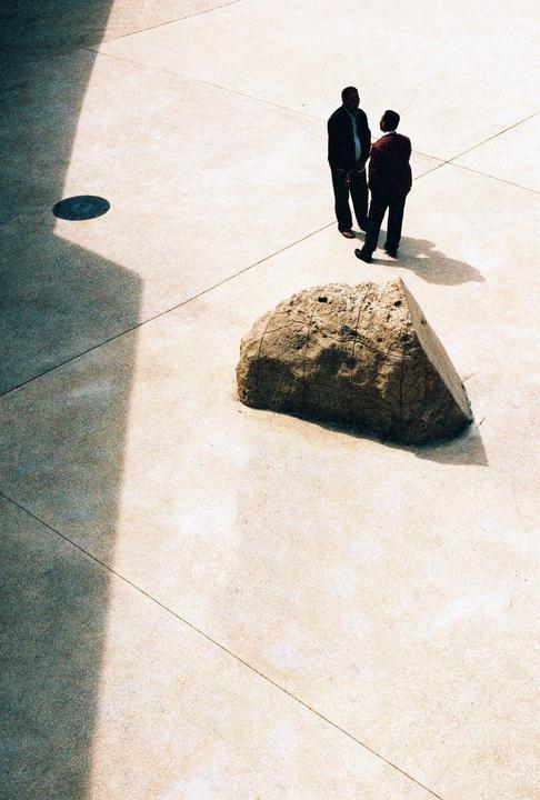 Two men stand on a light-colored paved surface next to a large rock, with shadows cast across the ground.