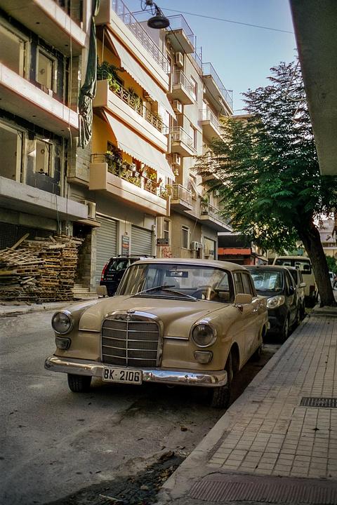 A vintage beige car is parked on a street next to a sidewalk and buildings.