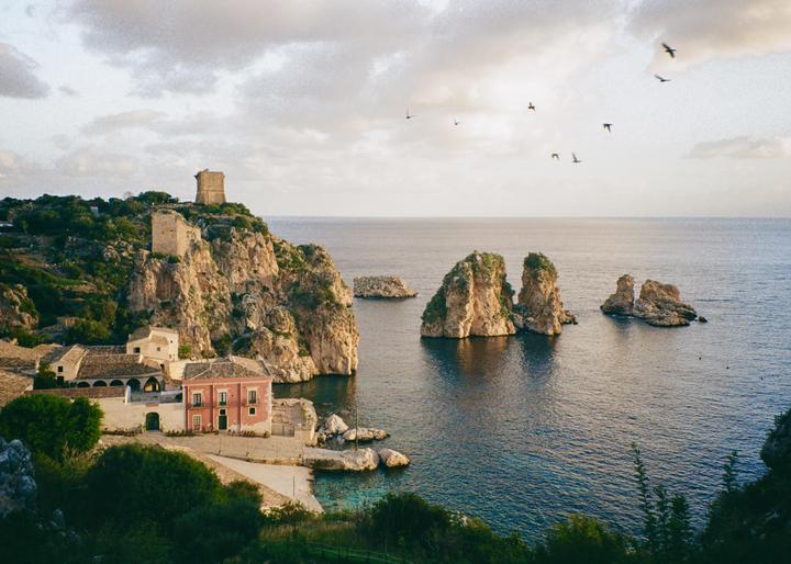 A small village with a pink building and a tower on a cliff overlooks the sea with several rock formations and birds flying in the sky.