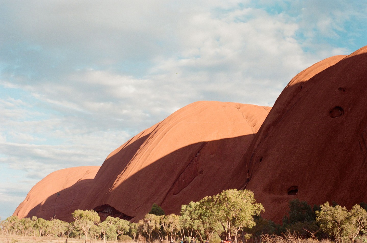 A large red rock formation with shadows cast across its surface stands under a partly cloudy sky, with green trees at its base.