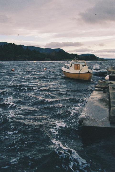 A small boat is docked next to a concrete pier on a choppy body of water with forested hills in the background under a cloudy sky.