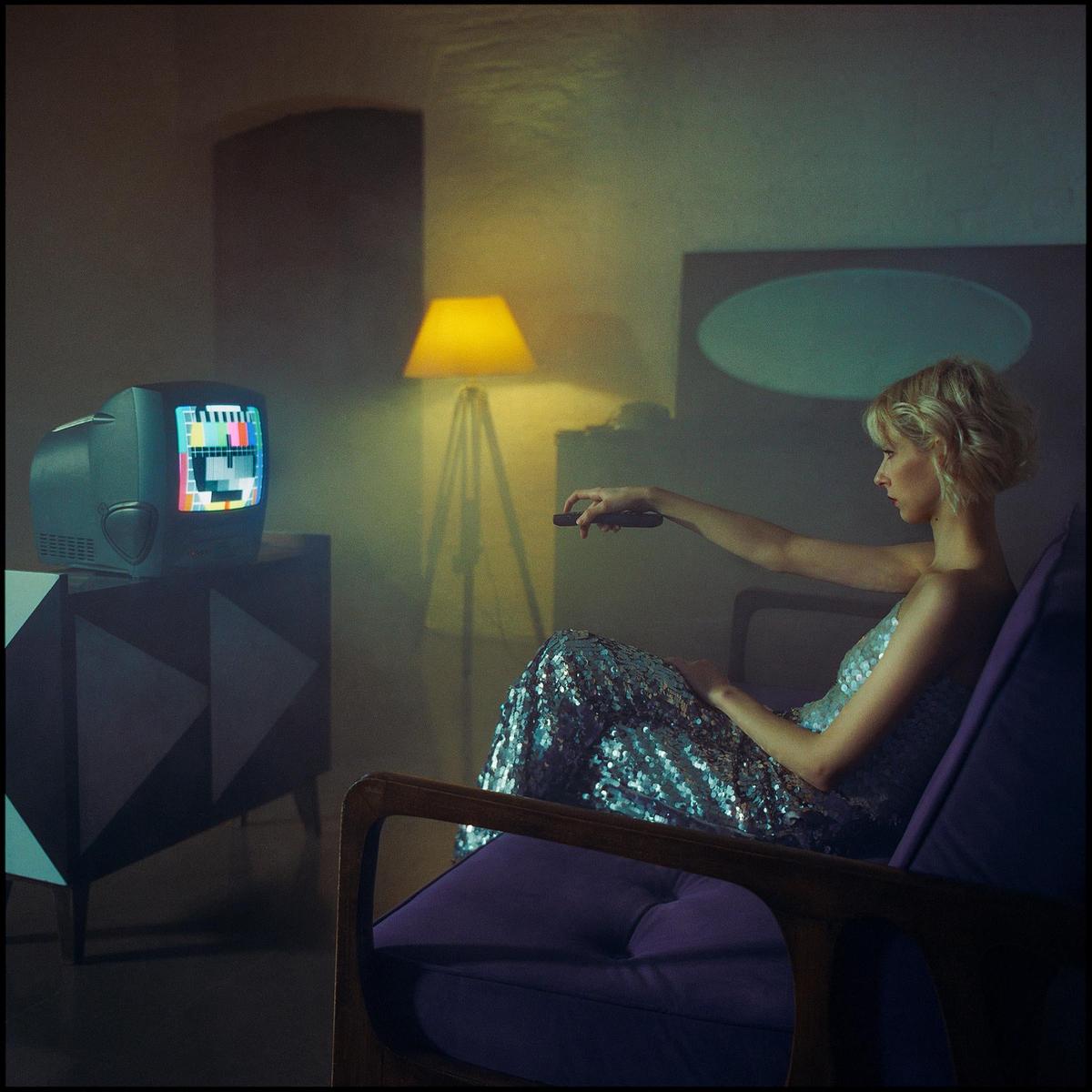 A woman in a sequin dress sits in a chair and points a remote control at a television displaying static.