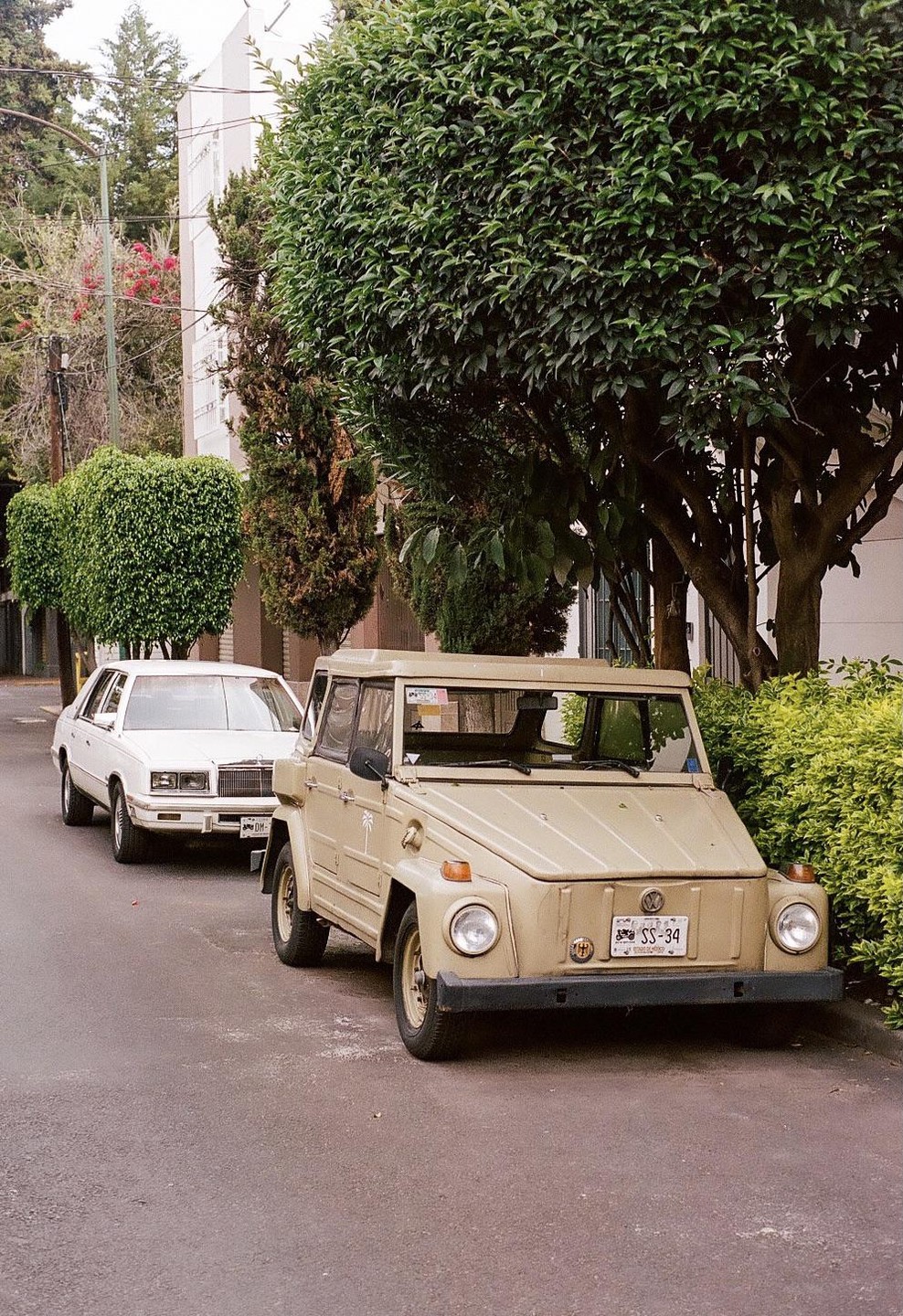 A beige Volkswagen Thing is parked on a street in front of a white car and lush green trees.