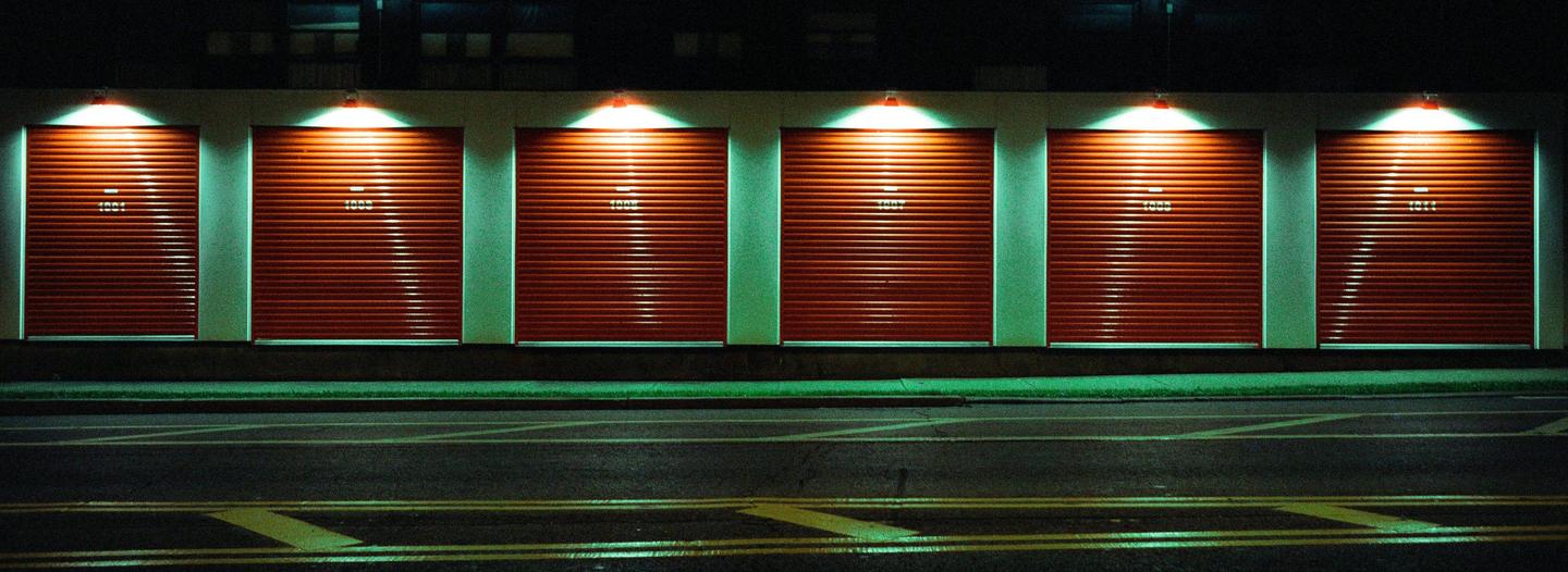 Six red garage doors with numbers are illuminated by overhead lights at night, with a street in the foreground.
