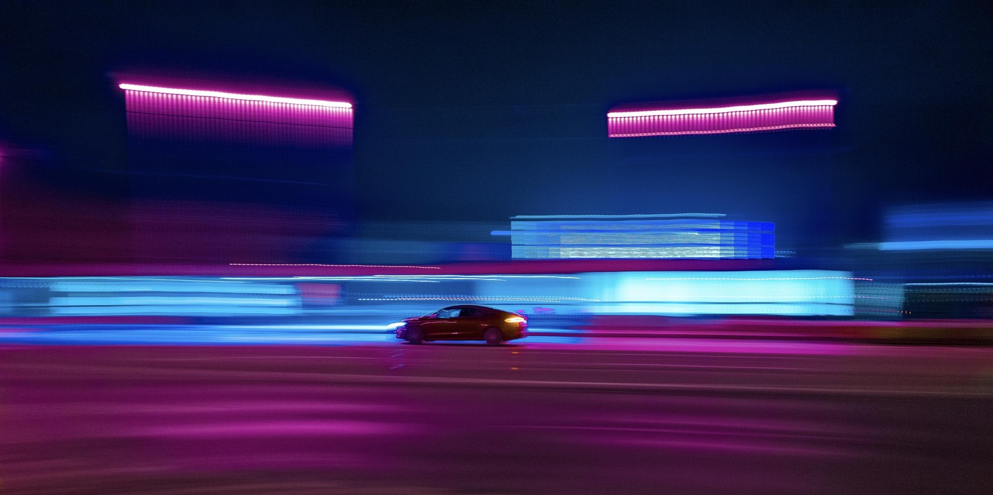 A car drives on a street at night with blurred neon lights in the background.
