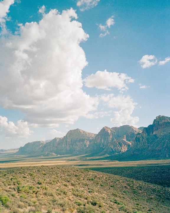 A vast desert landscape stretches towards a mountain range under a bright blue sky with fluffy white clouds.