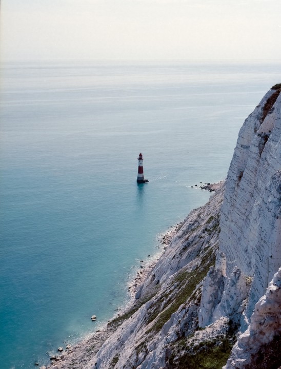 A red and white striped lighthouse stands in the blue sea next to a white cliff.