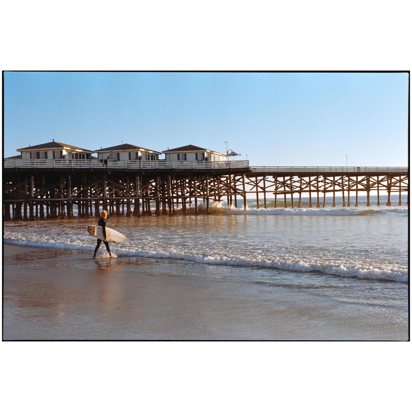 A surfer walks along the shore with a surfboard in front of a pier with buildings on it.