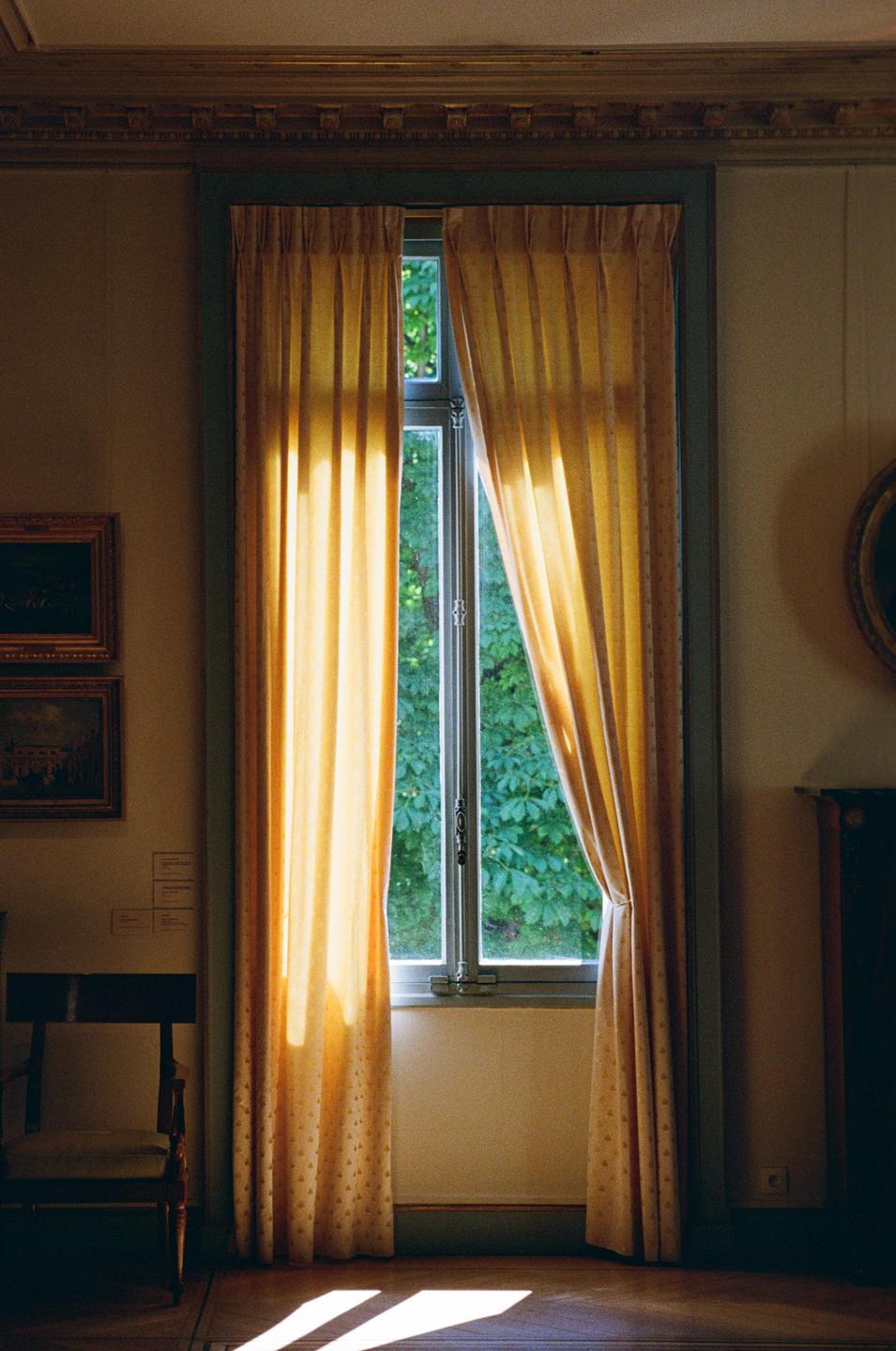 Sunlight streams through a window with yellow curtains, illuminating a room with a chair and framed artwork.
