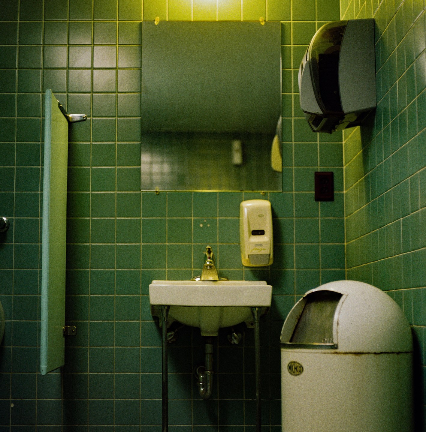A public restroom features green tiled walls, a sink, a mirror, a soap dispenser, a paper towel dispenser, and a trash can.