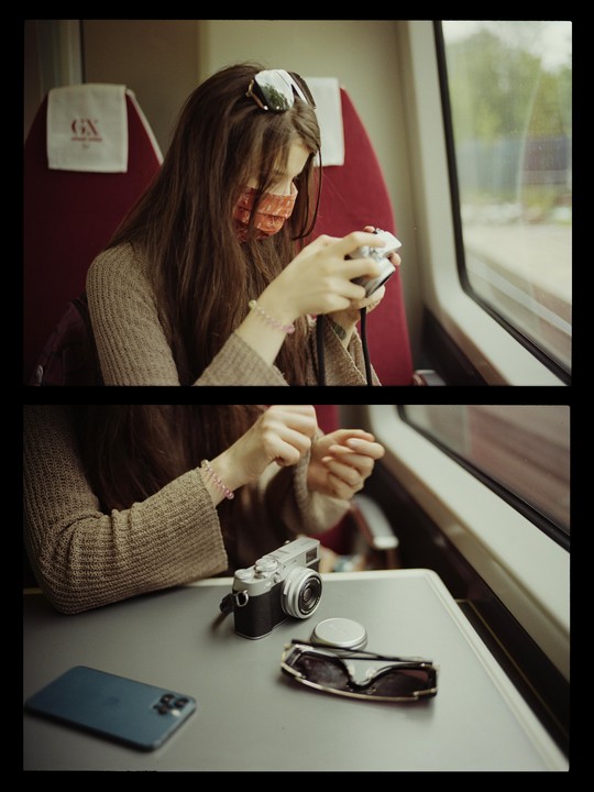 A woman wearing a face mask and sunglasses on her head looks at a camera while sitting on a train.