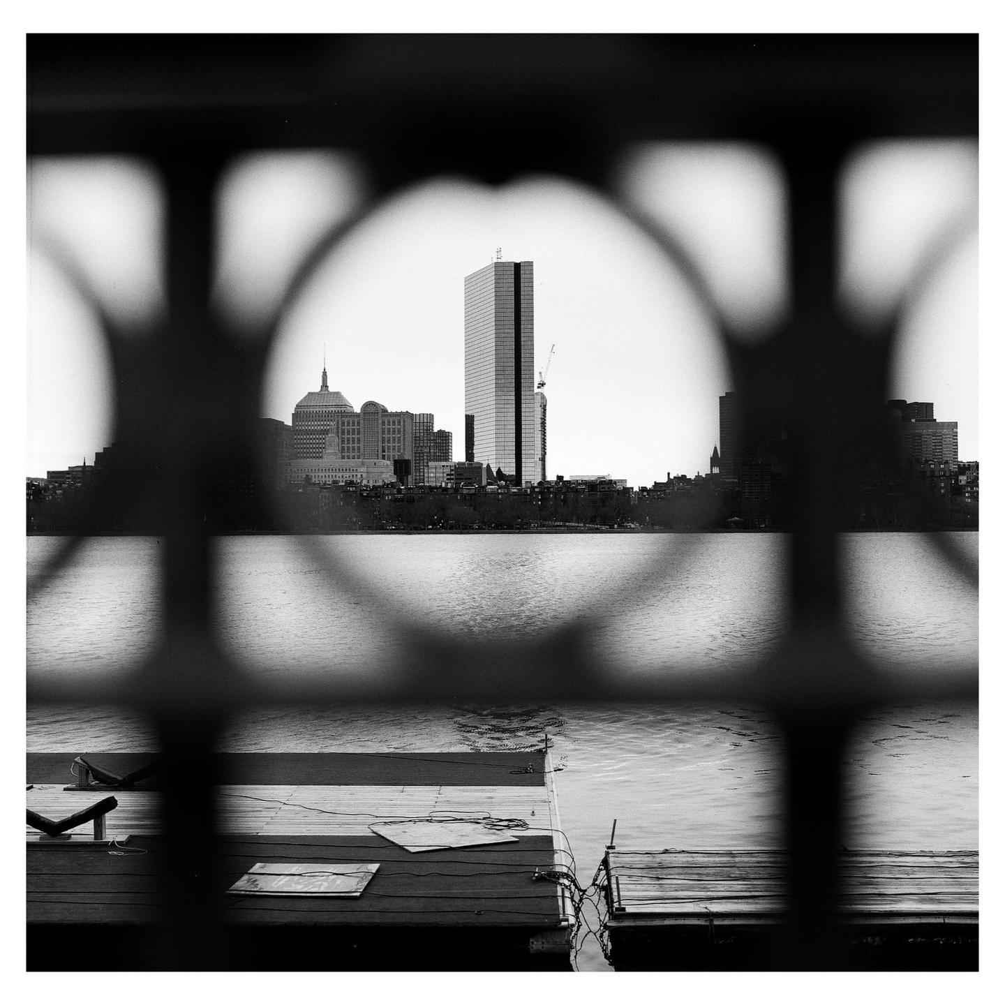A cityscape with a tall building is visible through a circular opening in a fence, with water and a dock in the foreground.