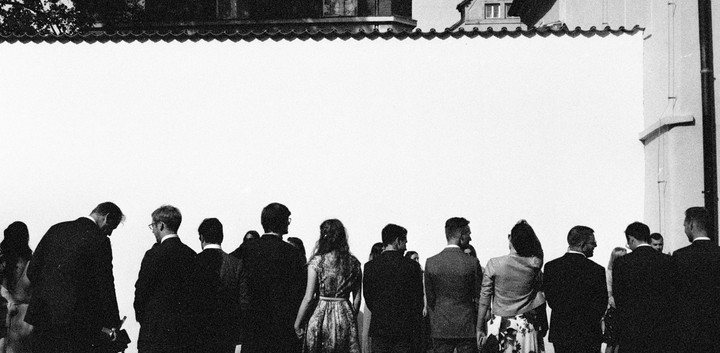 A group of people stand with their backs to the viewer, looking at a large white wall with a building visible above it.