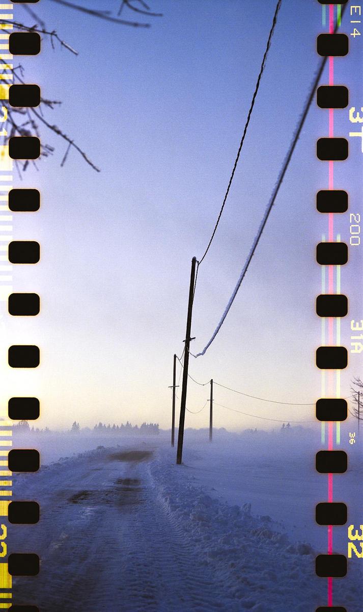 A snow-covered road with telephone poles stretches into the hazy distance under a pale sky.