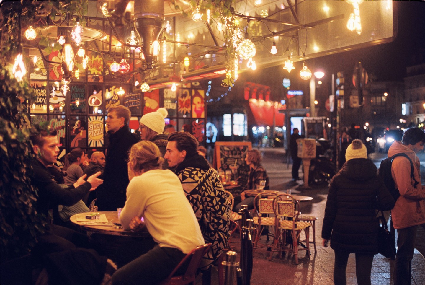 People sit and stand outside a bistro at night, illuminated by many small lights.