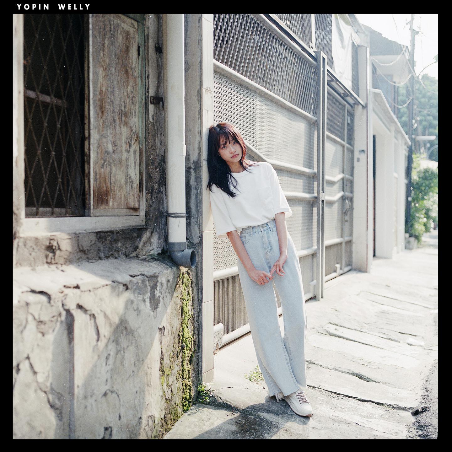 A woman with long dark hair leans against a wall on a sunny street.