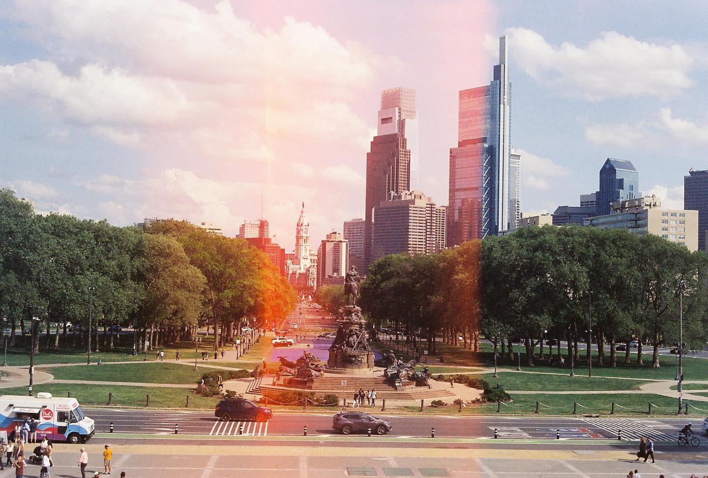 A wide view of a city park with a monument in the center, surrounded by trees and buildings under a cloudy sky.