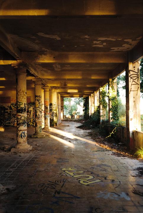 Sunlight streams through a long, abandoned, graffiti-covered hallway with pillars and overgrown ivy.
